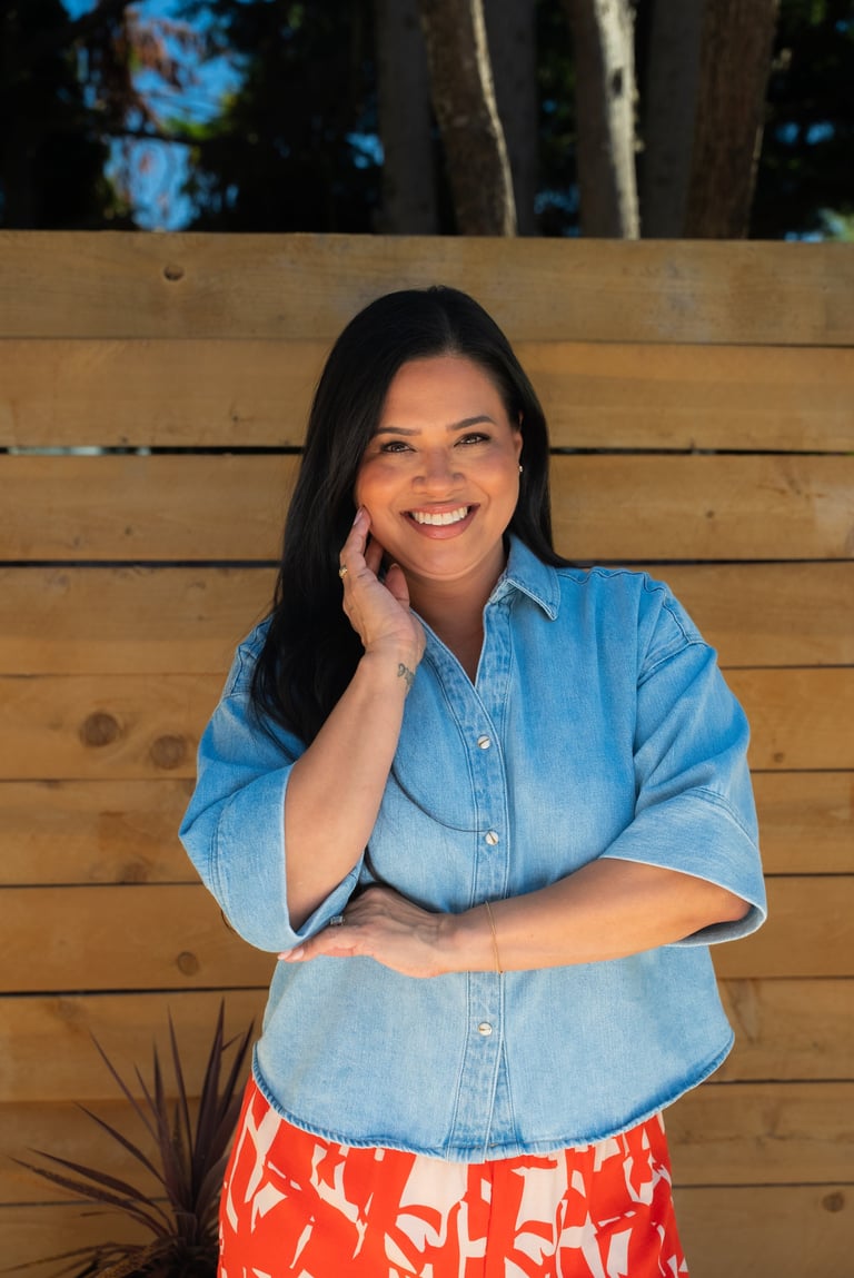 Woman smiling at camera wearing light blue denim shirt and red patterned skirt, leaning against wooden wall