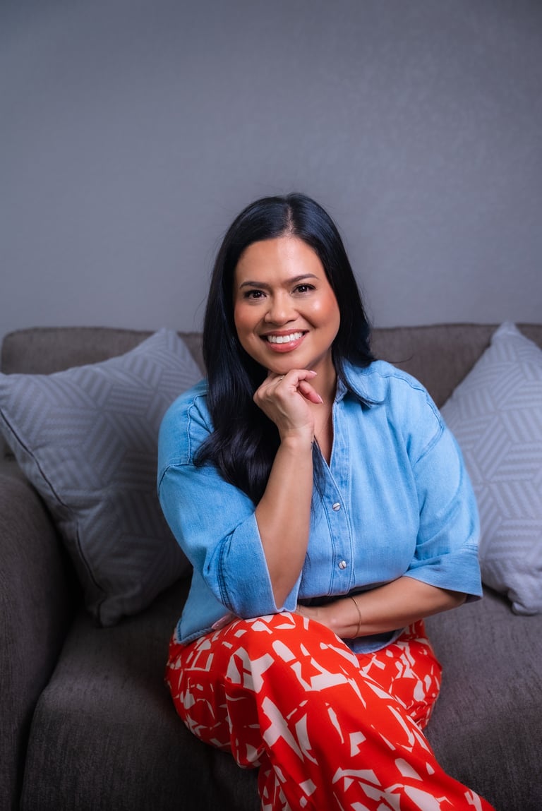 Woman with long dark hair smiling at camera, wearing light blue shirt and red patterned pants, sitting on gray couch